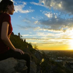 Woman sits on desert hillside overlooking a sunset in the city