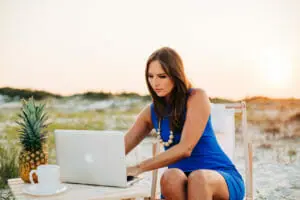 Woman with brown hair wearing blue dress works on laptop at beach
