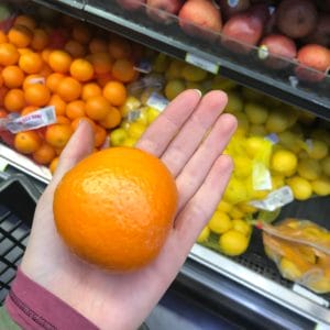 Orange being held on the palm of a hand in a store produce section.