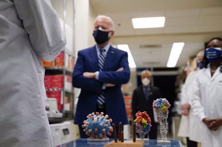 Joe Biden listening to a researcher talk, in a room of other researchers, all wearing masks, a COVID-19 virus model on a stand in the foreground