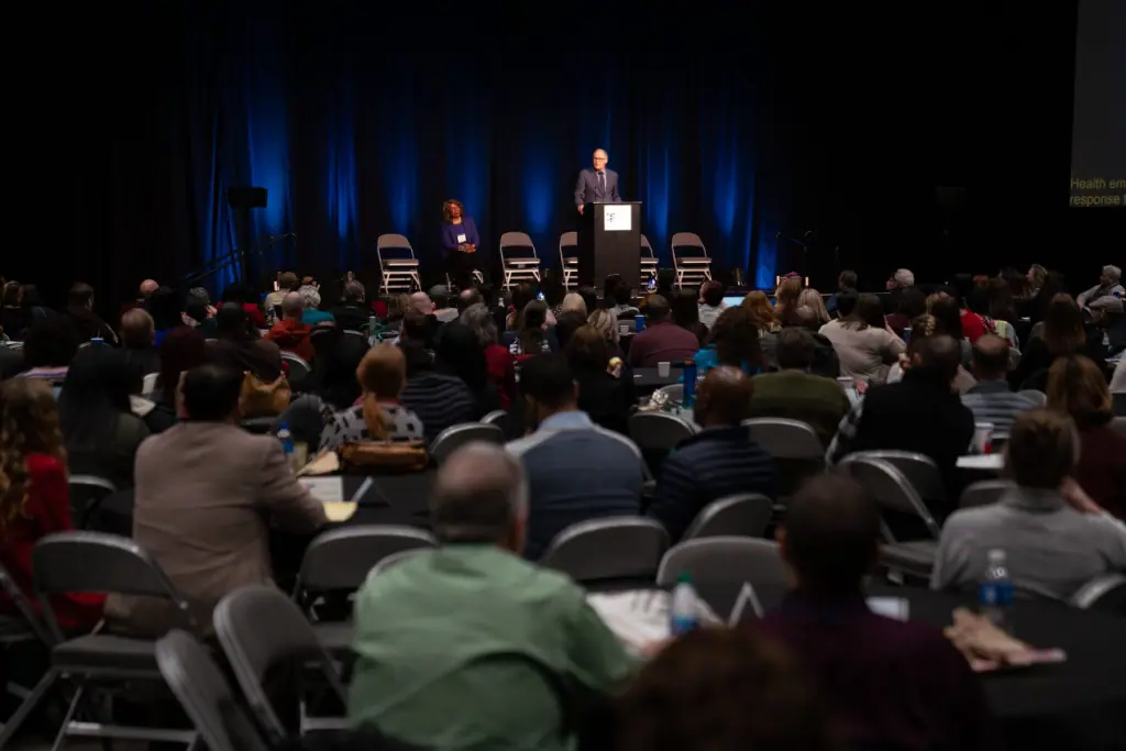 Wide-angle view of a conference setting with a speaker at a podium addressing a large audience. The speaker stands under focused stage lighting with blue accents, while the audience sits at round tables, attentively listening. A seated panelist is visible on stage, and the atmosphere is formal and professional.