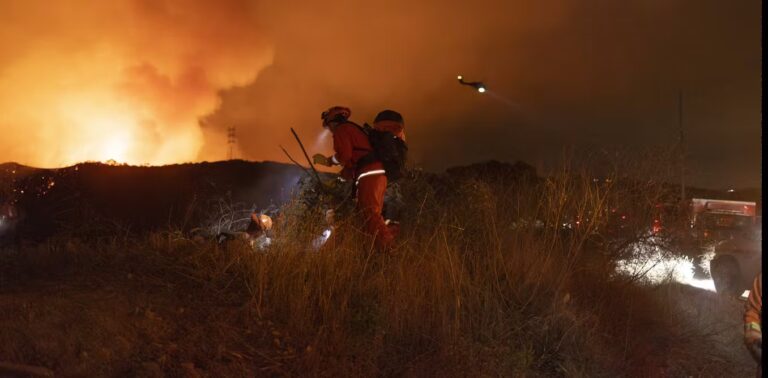 Firefighters battling a blaze in a smoky field at night, with intense flames and thick smoke filling the air. A helicopter is visible in the background, silhouetted against the dark, smoke-filled sky, assisting in the firefighting efforts.