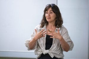 Pictured is Athena Aktipis, a person with long, dark hair wearing sunglasses on their head and dressed in a light-colored, loose-fitting jacket over a black top. They are standing in front of a whiteboard and gesturing with both hands raised and fingers slightly spread, as if emphasizing a point.