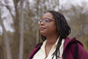 A person with long locs and glasses looks thoughtfully into the distance outdoors. They are wearing a maroon jacket over a light yellow shirt, with a soft, natural background of trees slightly out of focus.